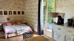 A bedroom with a stone wall, a bed with a patterned bedspread, a dresser with a lamp, and a window with a view of greenery. - Gîte "Ile d'Aix" with shared 7x7m swimming pool, 20 minutes from the beach (Saint-Coutant-Le-Grand)