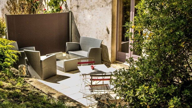 A patio area with wicker furniture, a glass table, and a view of a building with a door and window. - Gîte "Ile d'Aix" with shared 7x7m swimming pool, 20 minutes from the beach (Saint-Coutant-Le-Grand)