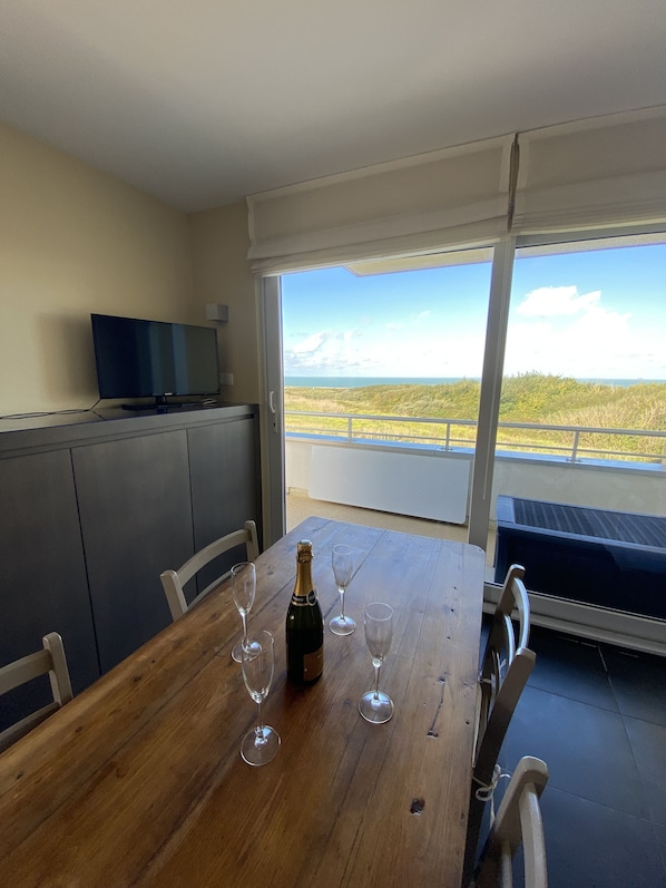 A dining table with a bottle of champagne and glasses, a television on a cabinet, and a balcony with a view of the beach.