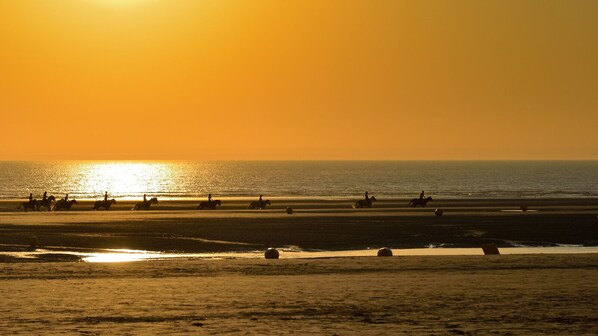 Una playa con gente montando a caballo, un atardecer y el océano.