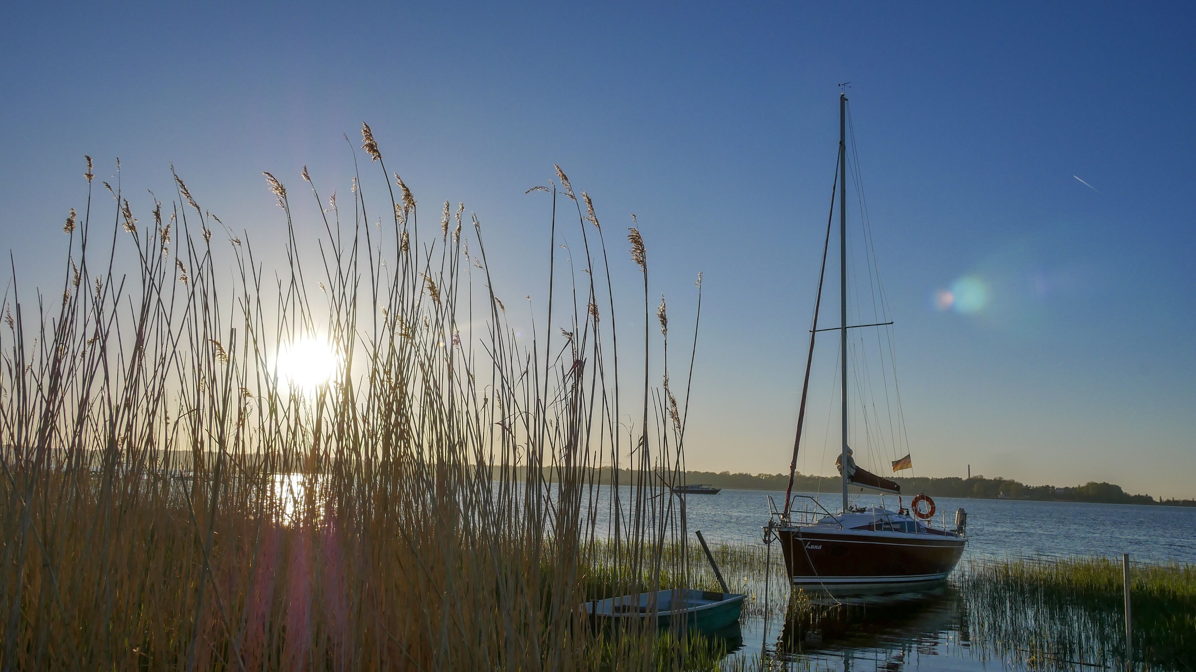 Un lac paisible, avec de hautes herbes au premier plan et un voilier amarré sur la droite.