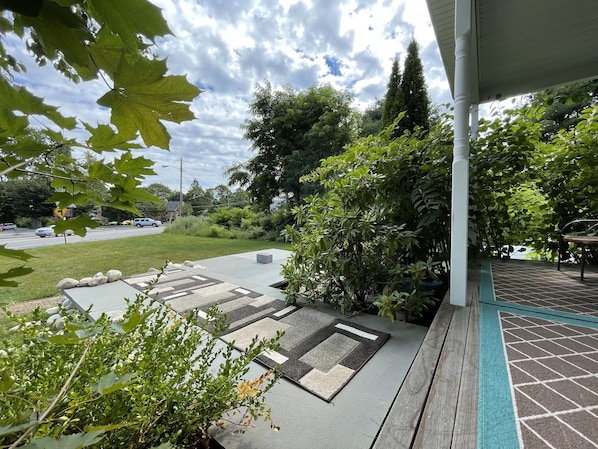 A patio area with a patterned rug, a wooden bench, and a view of a road with cars and greenery in the background.