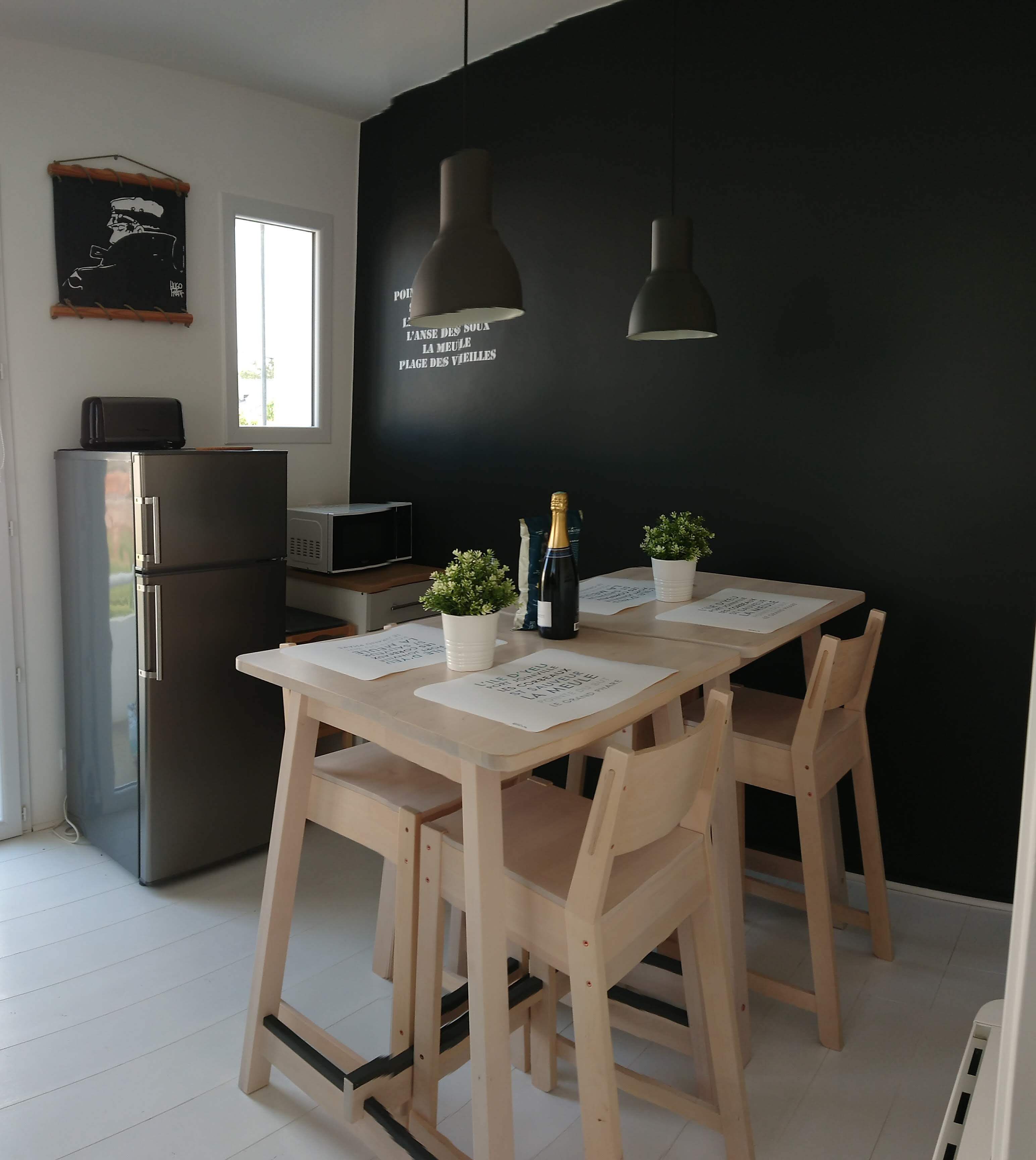 A modern kitchen with a black accent wall, wooden dining table, and chairs. There is a refrigerator, a microwave, and a small potted plant on the table.