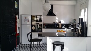Una cocina moderna con piso a cuadros blanco y negro, un refrigerador blanco, una estufa y un lavamanos. Hay dos taburetes negros y un tazón de fruta en la encimera.