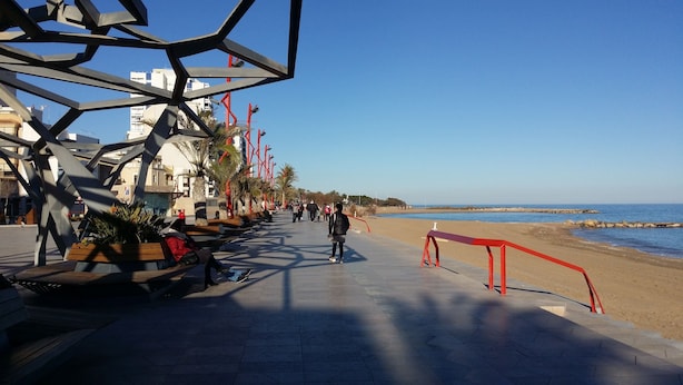 Un paseo marítimo con bancos, palmeras y una playa al fondo.