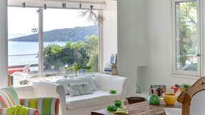 A bright living room with a white sofa, colorful striped armchair, and a wooden chair with a cushion. A wooden coffee table and a window with a view of a body of water and greenery are visible. - Beatifull seaview / 100m from beach (Amaliapolis)