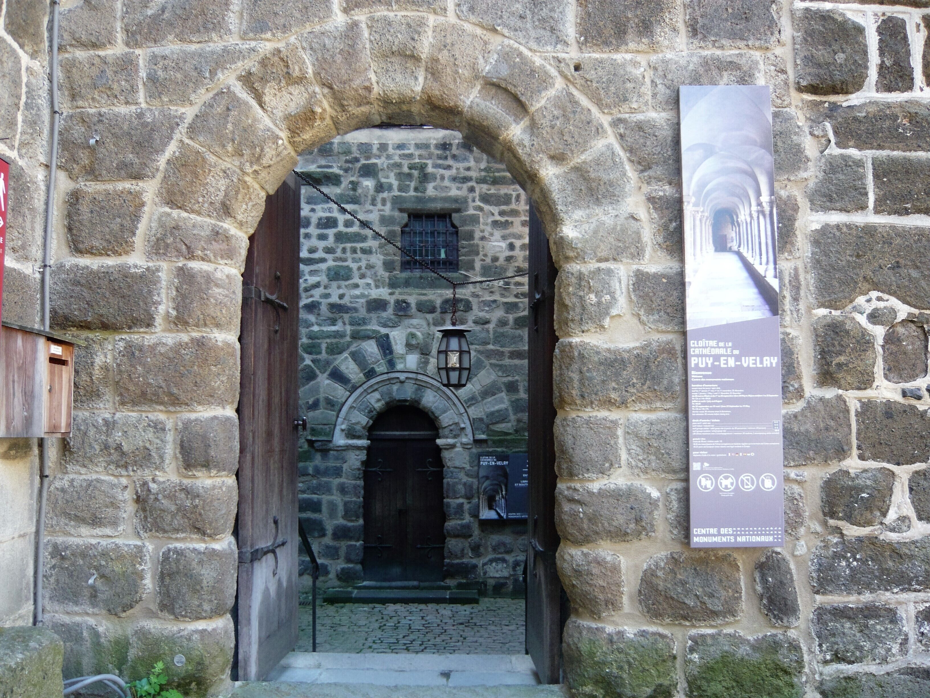 Un arc en pierre mène à un bâtiment doté d’une porte en bois et d’une lanterne.