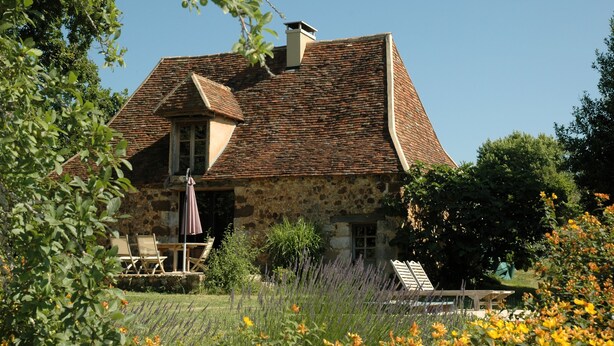 Ein Steinhaus mit geneigtem Dach, einem Schornstein und einem Garten voller Blumen und Pflanzen.