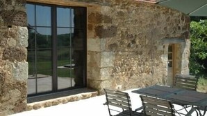 Ein Gebäude aus Stein mit einem Ziegeldach, einer großen Glasdoor und einer Terrasse mit Tisch und Stühlen.