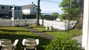 A white house with a white picket fence, a paved pathway, and two white chairs in the foreground.