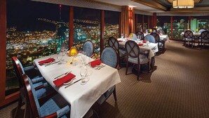 Un restaurant avec des tables dressées pour le service, de grandes fenêtres offrant une vue sur la ville et un sol recouvert de moquette.