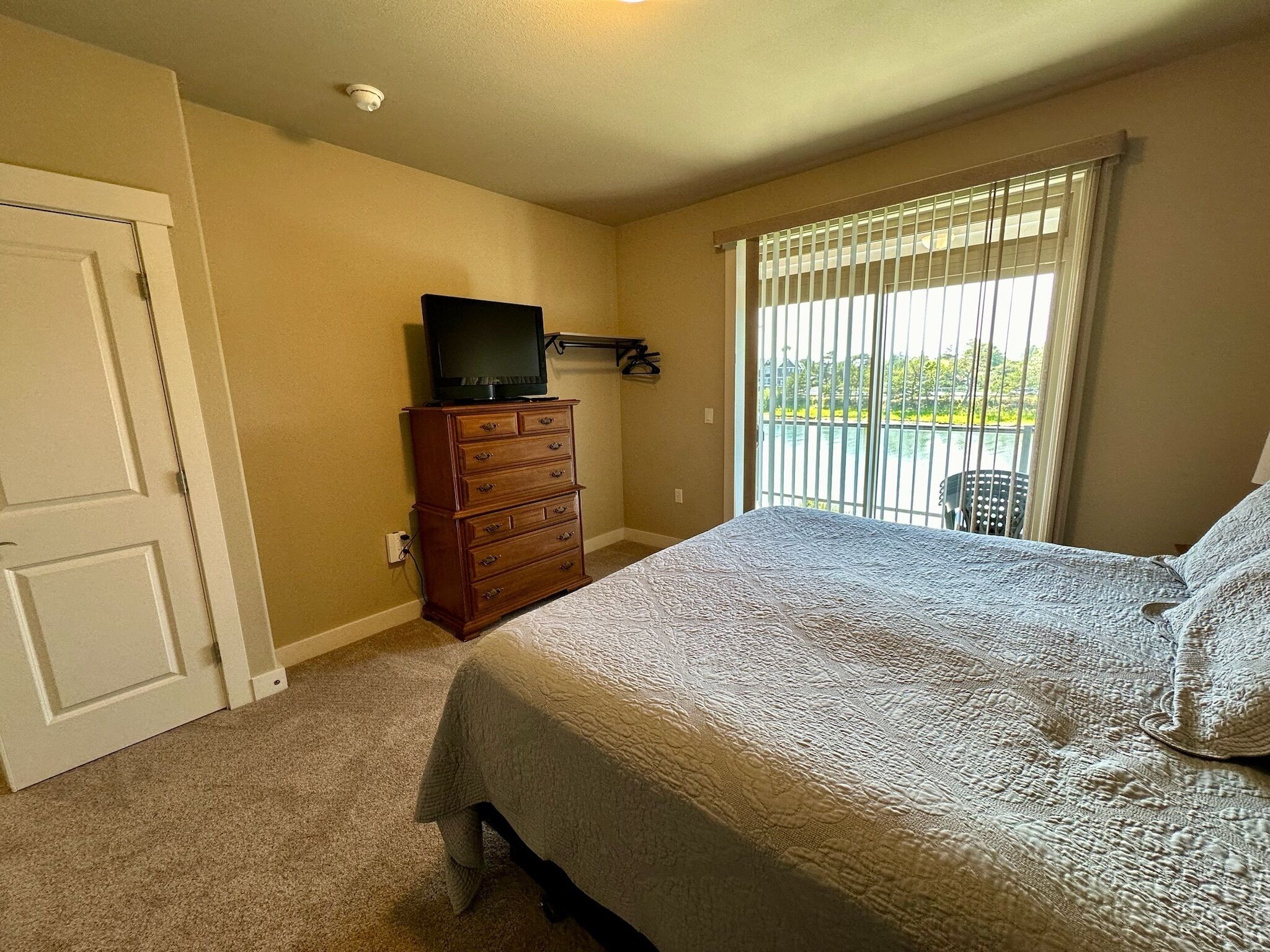 Un dormitorio con cama, cómoda, televisor encima y una ventana con persianas, además de una puerta.