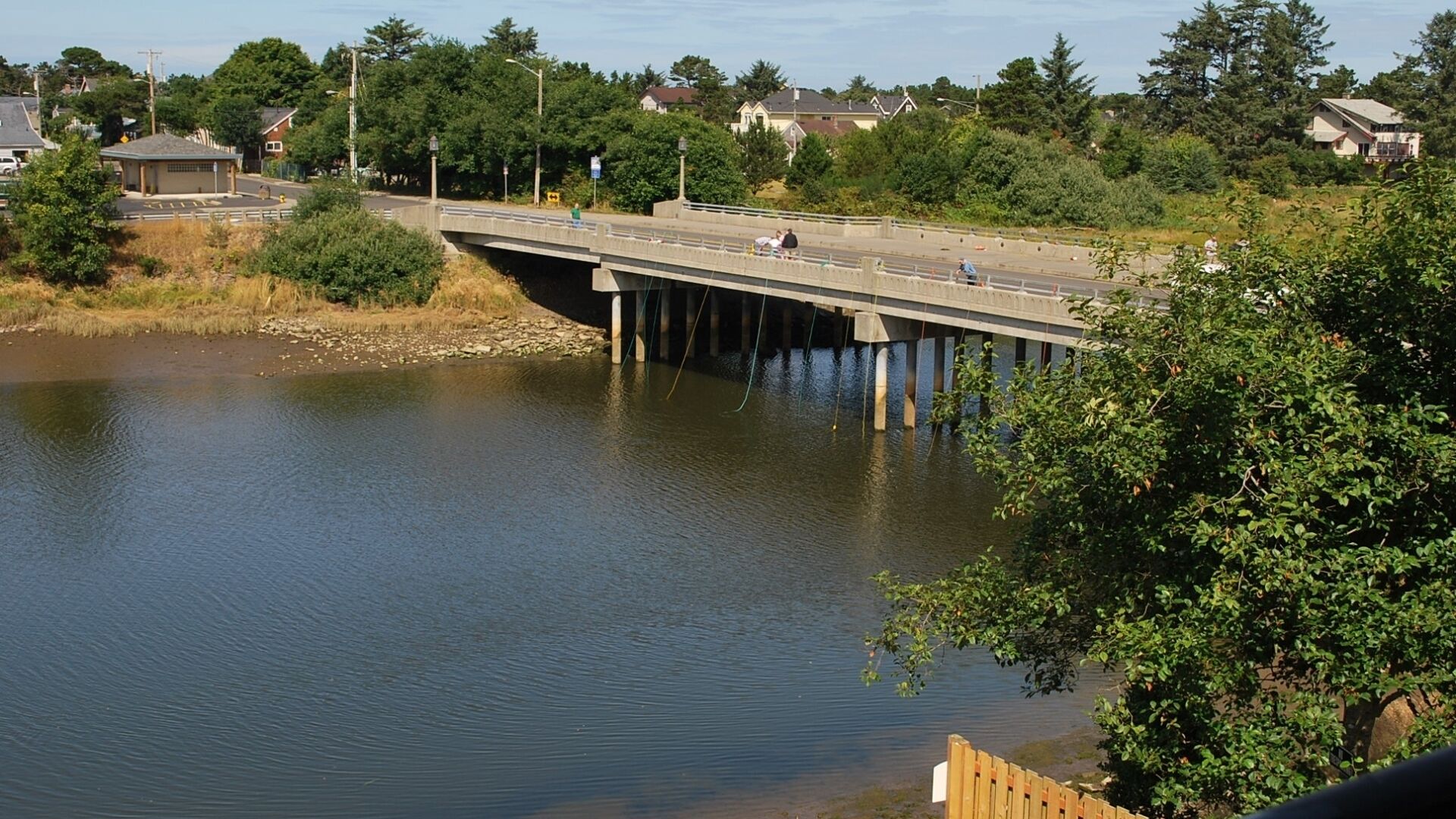 A concrete bridge over a calm river with a pedestrian walkway, surrounded by greenery and residential buildings.