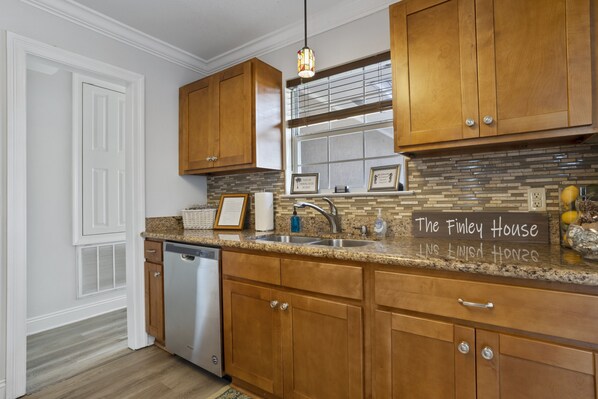 Una cocina con armarios de madera, encimera de granito, pileta de acero inoxidable y lavavajillas. Hay un cartel en la encimera que dice "The Finley House".