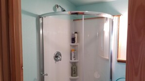 Una cabina de ducha con paredes de vidrio y puerta, un estante pequeño con artículos de aseo, y una silla azul en la esquina.