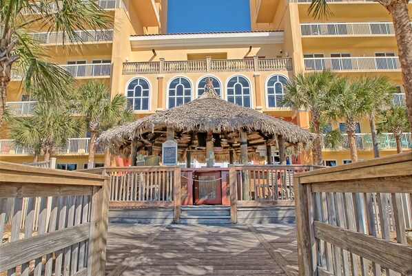 A wooden boardwalk leading to a building with a thatched roof structure, balconies, and palm trees.