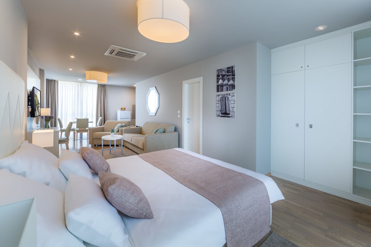 A modern hotel room with a large bed, a desk, a chair, a wardrobe, and a dining area.