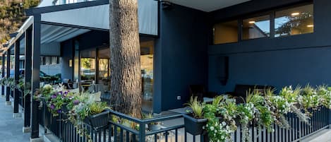 A modern building with a covered outdoor seating area, a tree, and potted plants along the railing.