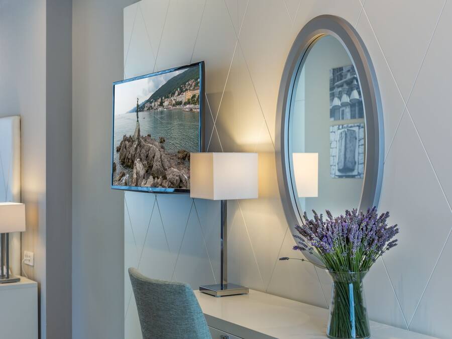Een moderne hotelkamer met een grote spiegel, een flatscreen televisie, een bedlamp, een stoel en een vaas met lavendelbloemen.