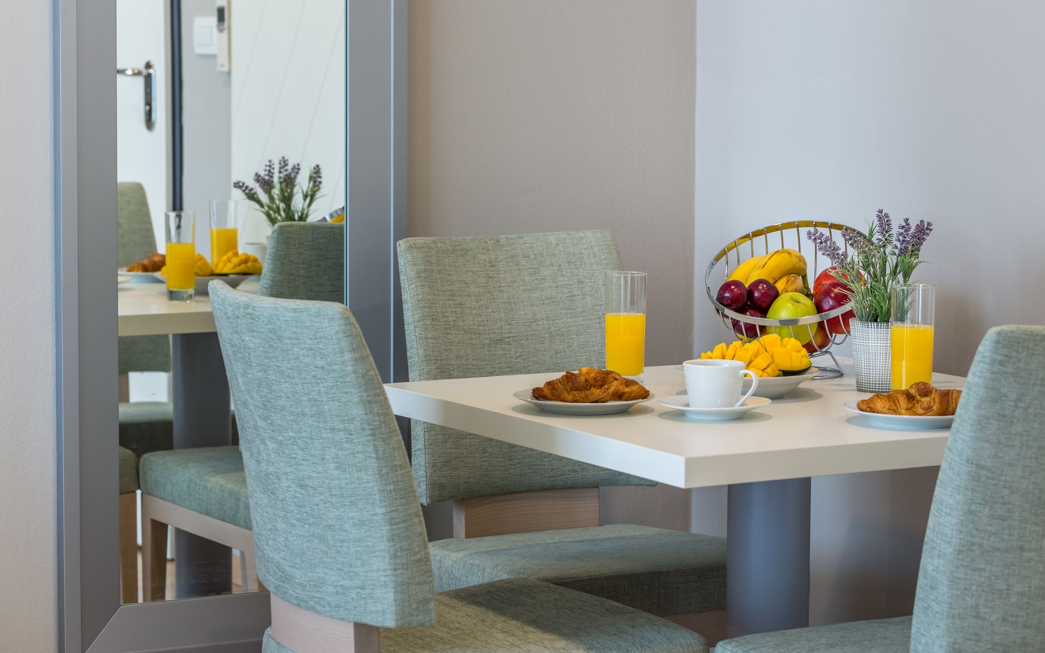 Une table mise pour le petit-déjeuner, avec des croissants, des fruits et du jus, entourée de chaises grises à revêtement rembourré.