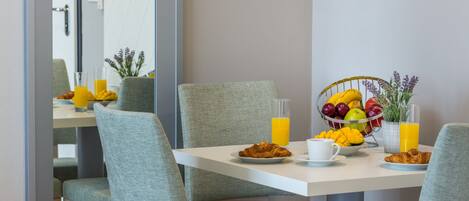 Une table mise pour le petit-déjeuner, avec des croissants, des fruits et du jus, entourée de chaises grises à revêtement rembourré.