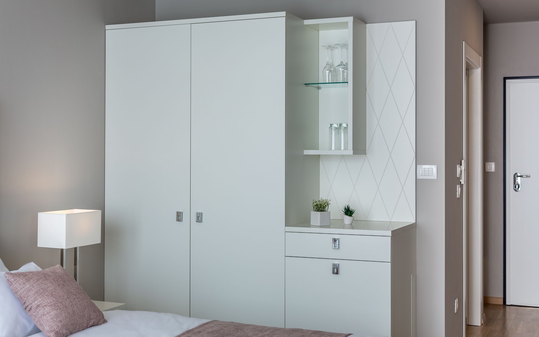A bedroom with a bed, a nightstand, a bedside lamp, a wardrobe, a mirror, and a small shelf with decorative items.