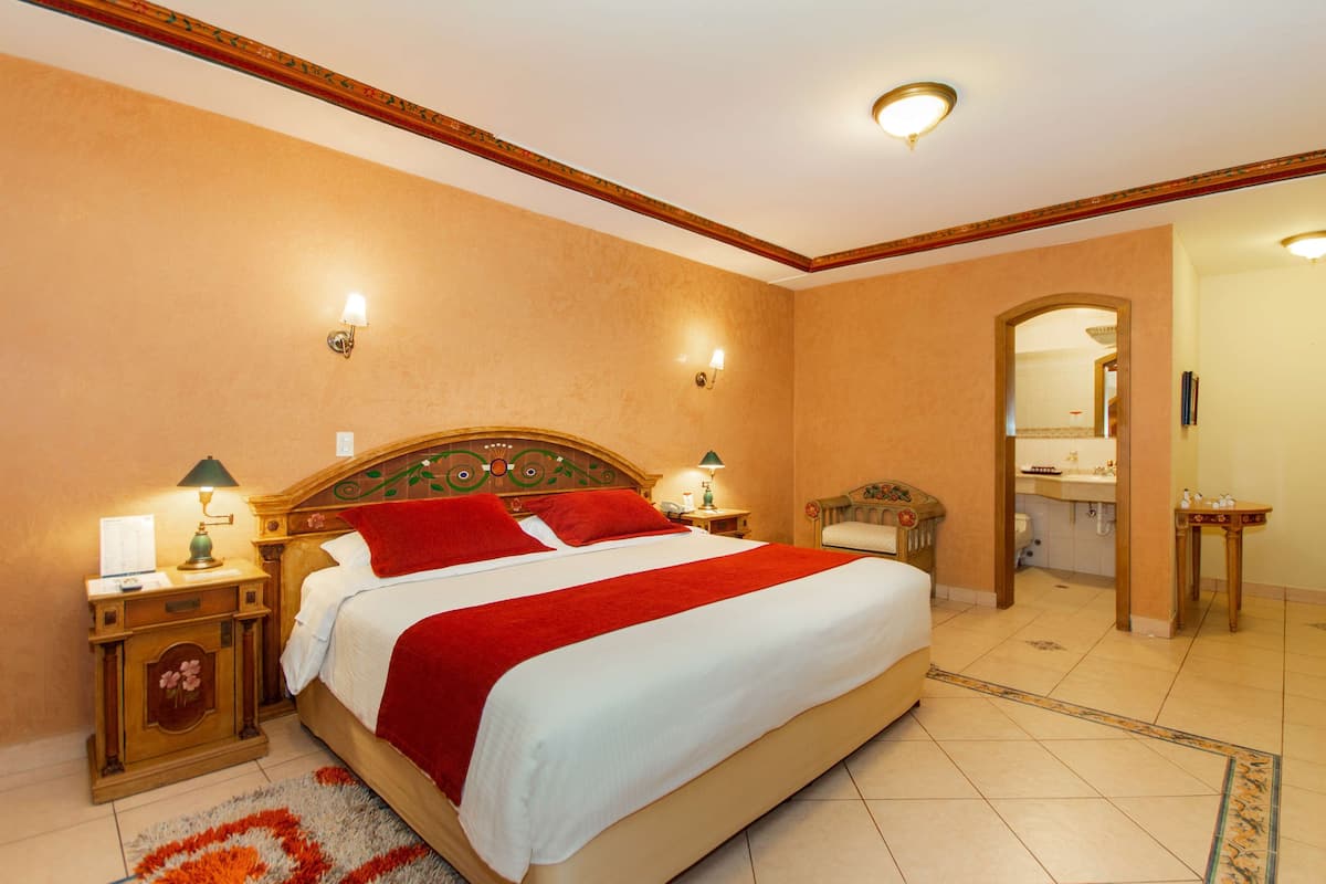 A hotel room with a large bed, two bedside tables with lamps, a wooden headboard, a bathroom visible through an archway, and a patterned rug on the floor.