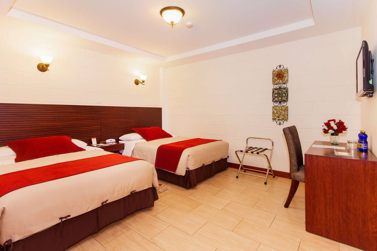 A hotel room with two beds, a desk, a chair, and a small trolley.