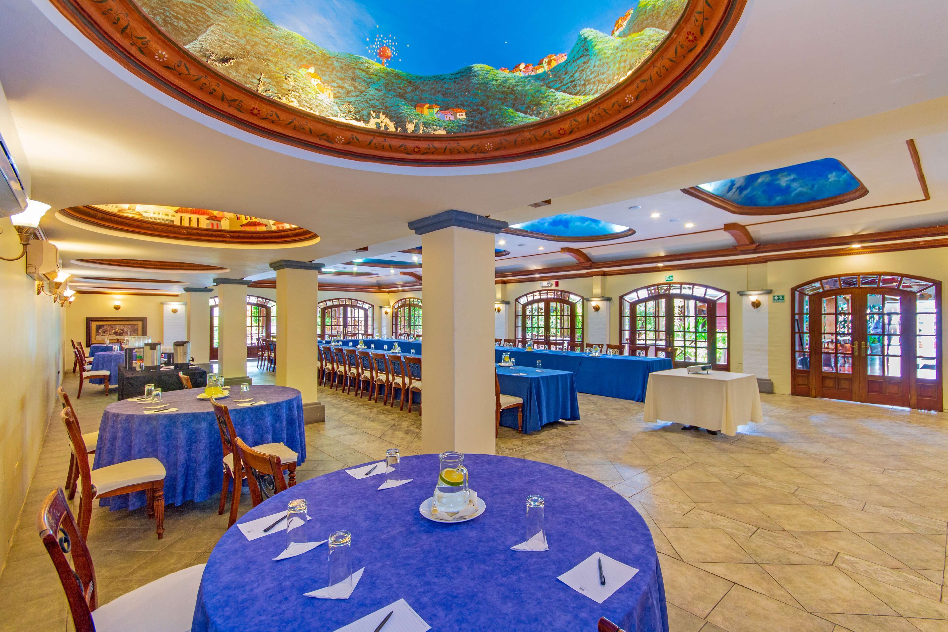 Une salle de banquet spacieuse, dotée de tables rondes, d’un plafond haut orné d’une fresque et de grandes fenêtres.