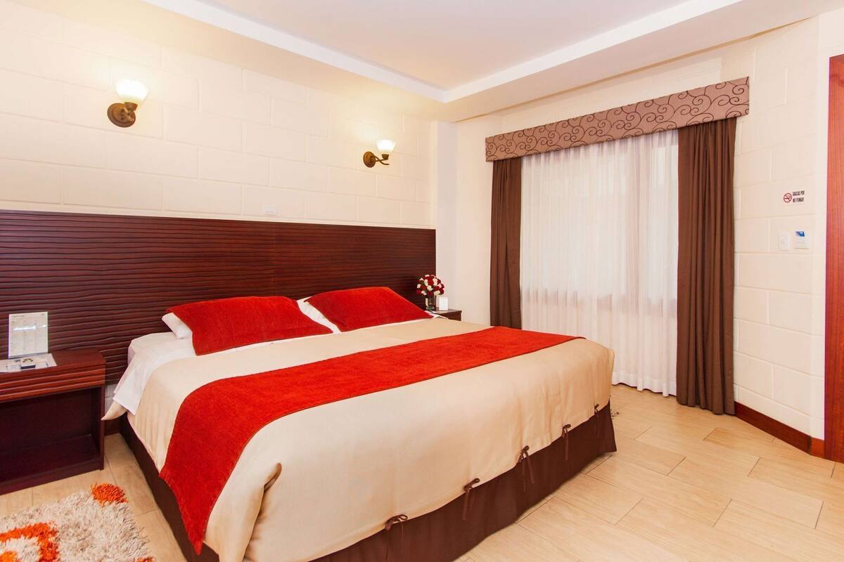 A hotel room with a large bed, two bedside tables, a nightstand, a wall-mounted lamp, a window with curtains, and a wooden floor.
