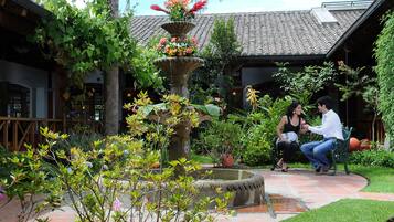 Un patio con una fuente de piedra, abundante vegetación y una estructura de techo con tejas.