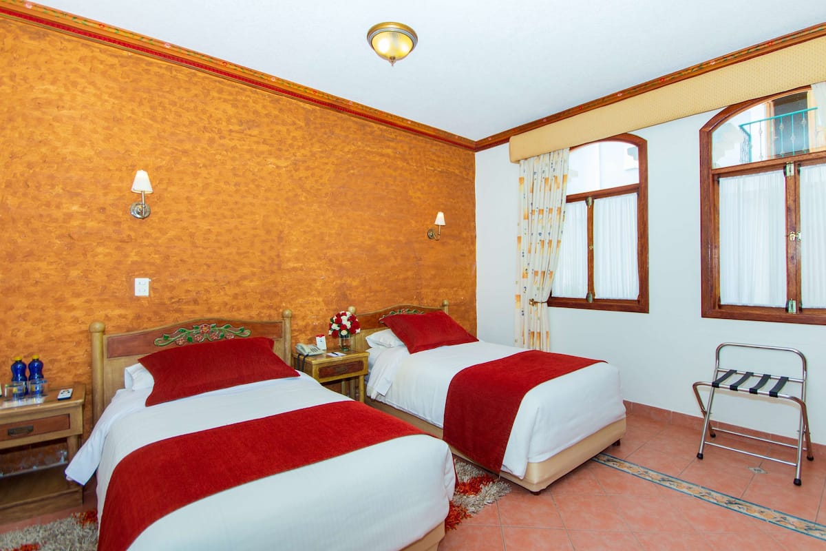 A hotel room with two beds, a wooden headboard, a nightstand with a lamp, a window with curtains, and a radiator.