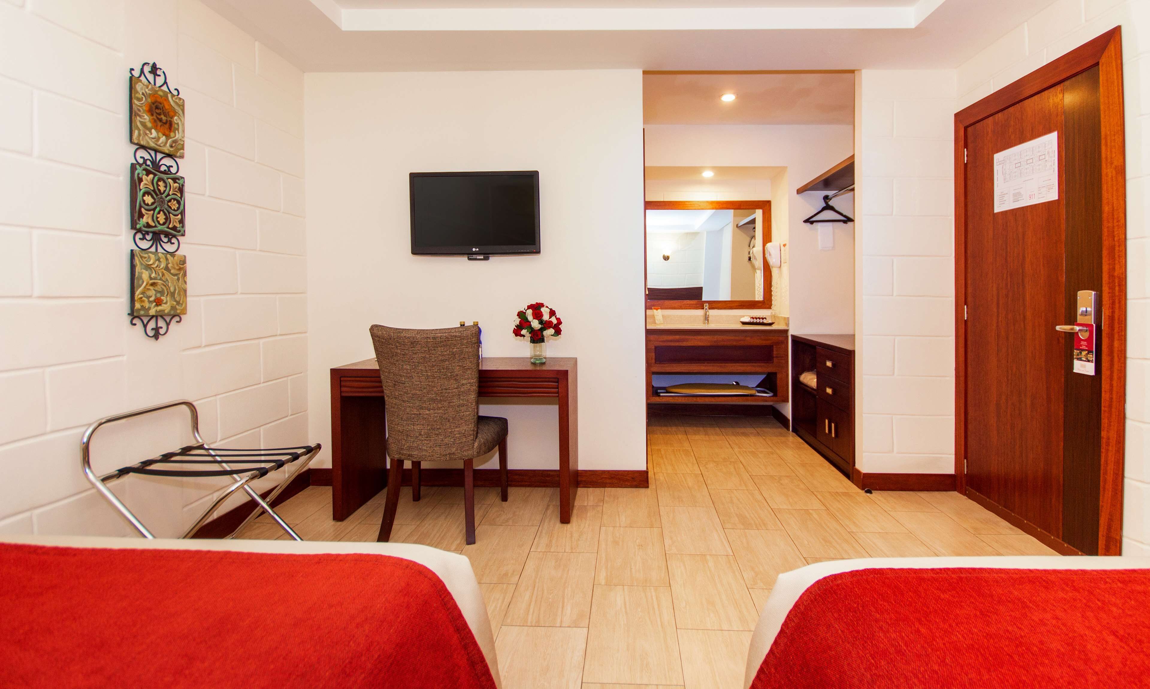 A hotel room with two beds, a desk with a chair, a TV, a mirror, and a bathroom visible through an open door.
