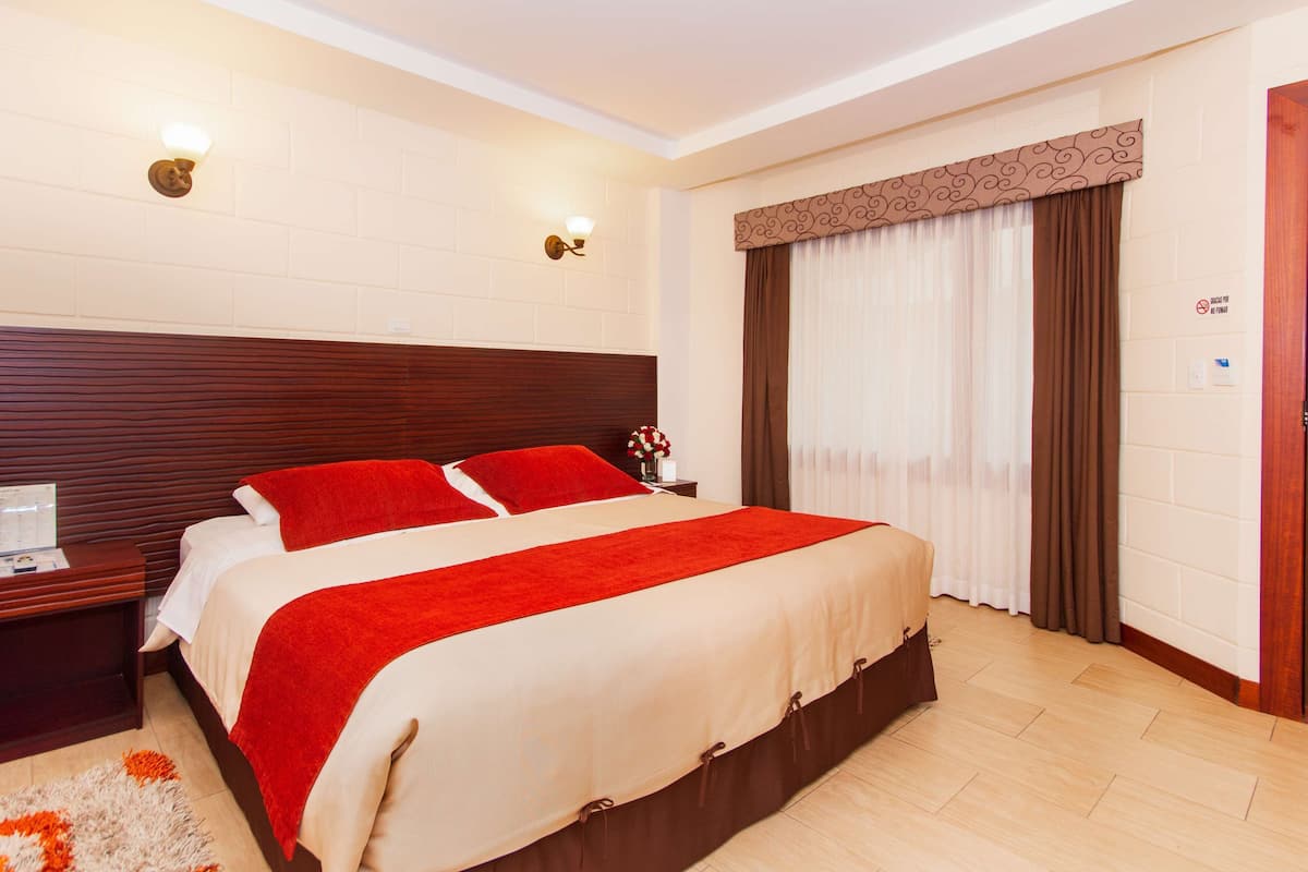 A hotel room with a large bed, a wooden headboard, a nightstand with a lamp, a window with curtains, and a door with a sign.
