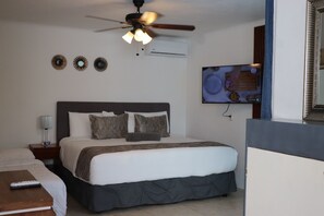Habitación de hotel con cama, mesita de noche con lámpara, ventilador de techo, decoración en la pared y televisor.