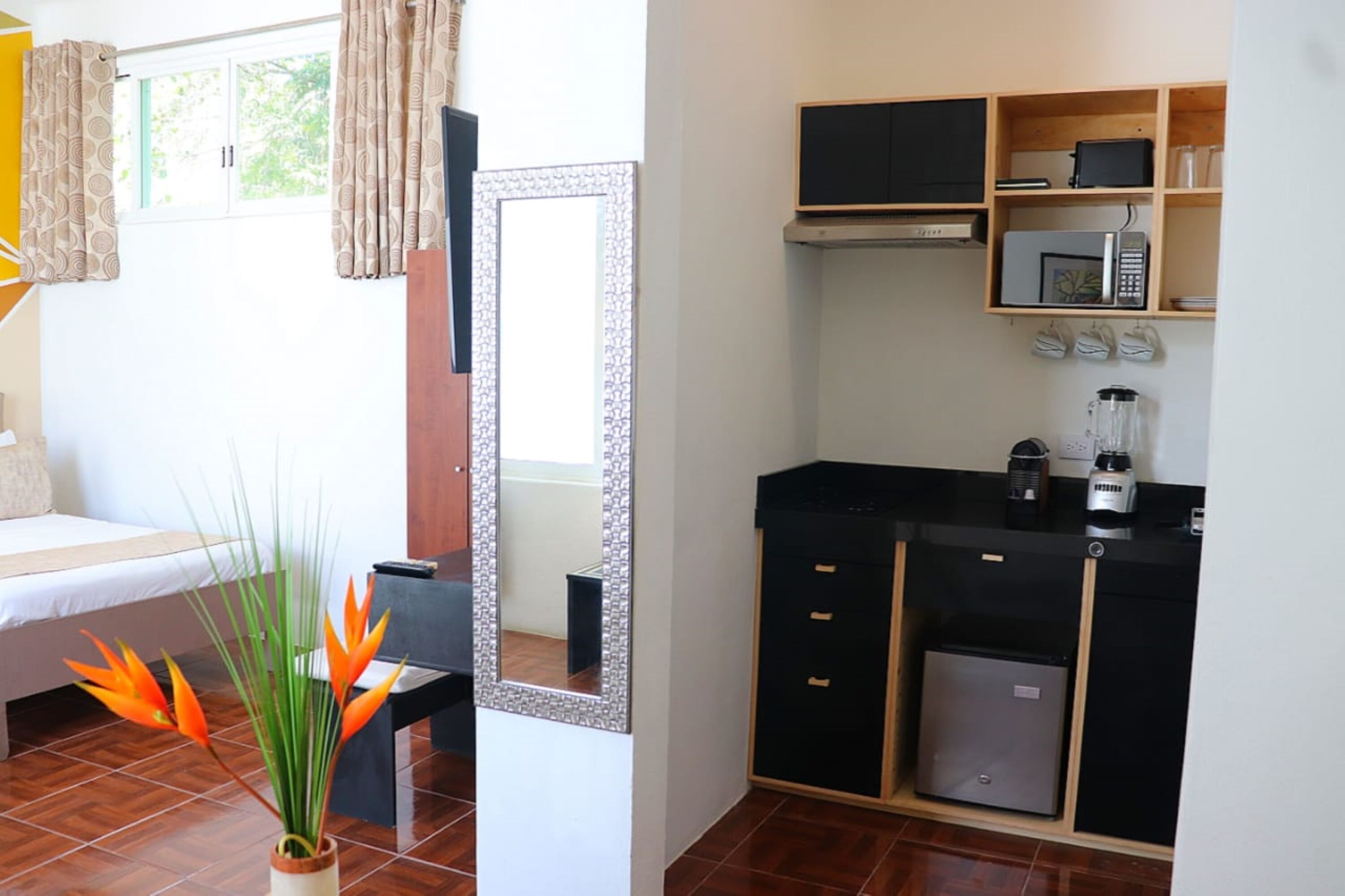 Una habitación con cama, un espejo, una mesita con una planta, una kitchenette con microondas y cafetera.