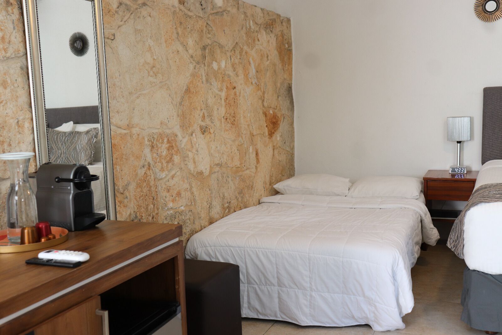 Una habitación de hotel con pared de piedra, un escritorio de madera con cafetera, cama con ropa blanca y mesita de noche con lámpara.