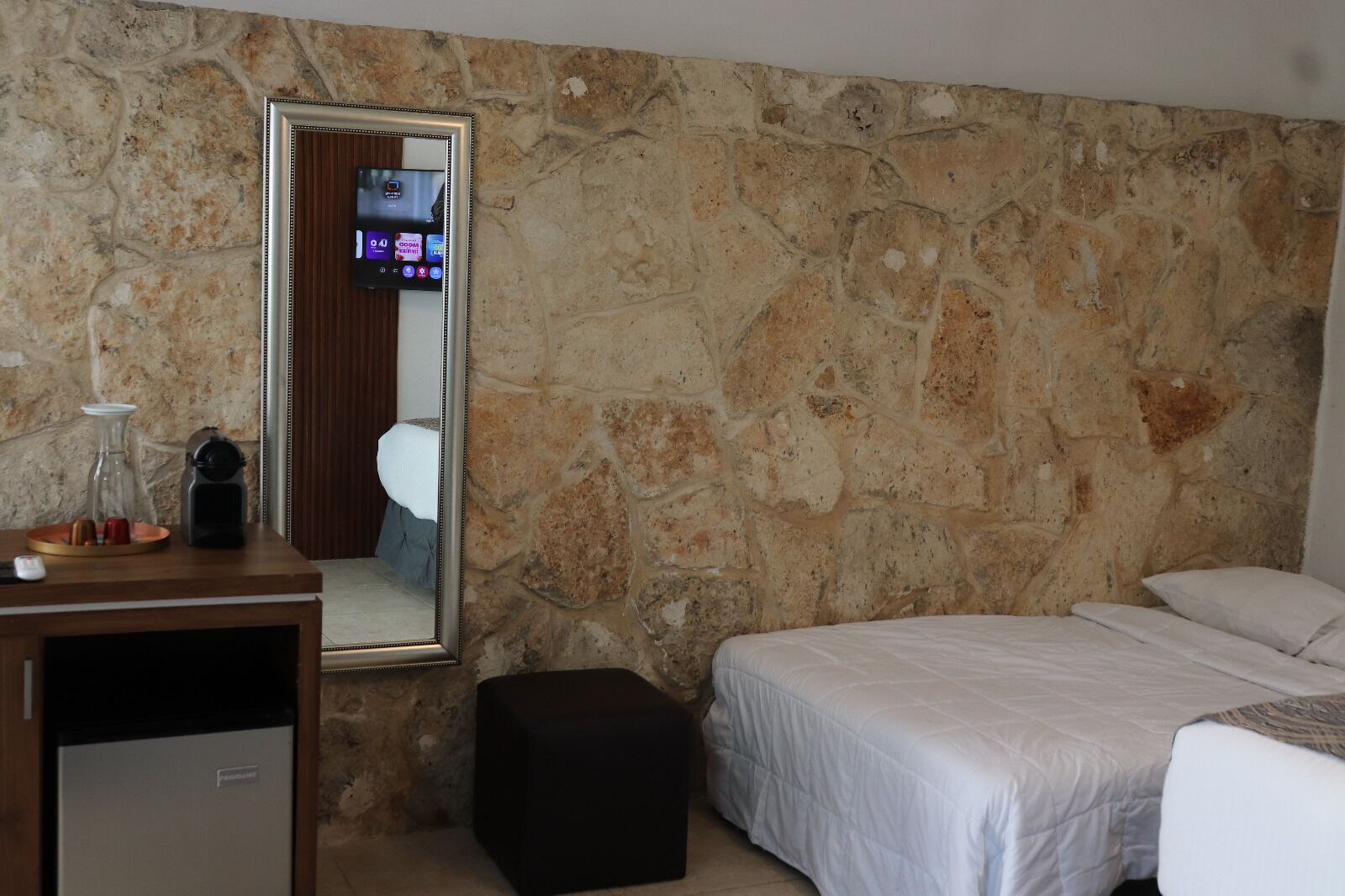 A bedroom with a stone wall, a bed, a bedside table, a mirror, and a television.