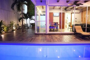 Un área de piscina interior moderna con un salón envuelto en vidrio, suelo de madera y ventiladores de techo.