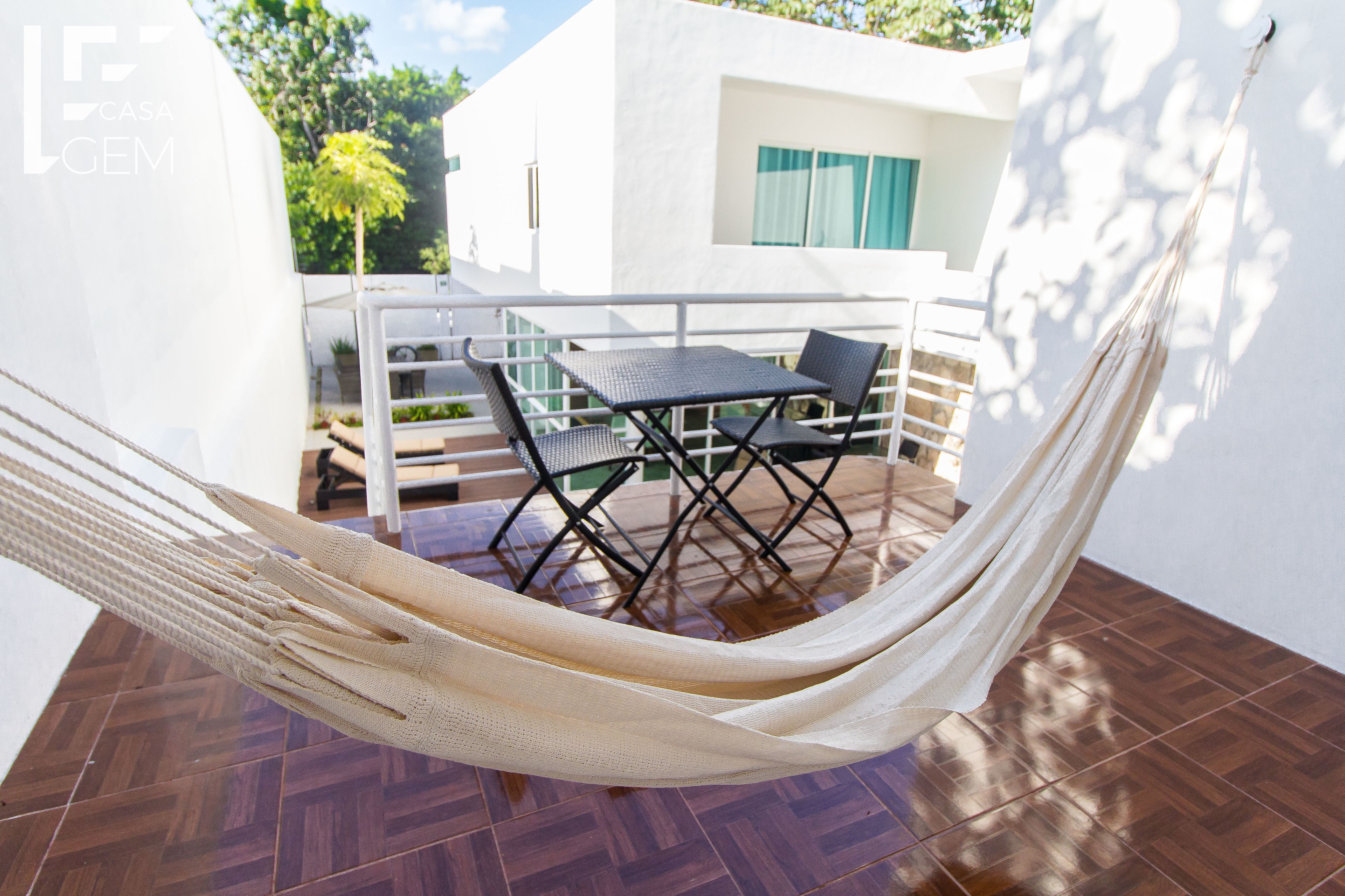 Een hangmat op een betegeld terras met een tafel en stoelen, een wit gebouw op de achtergrond en een boom.