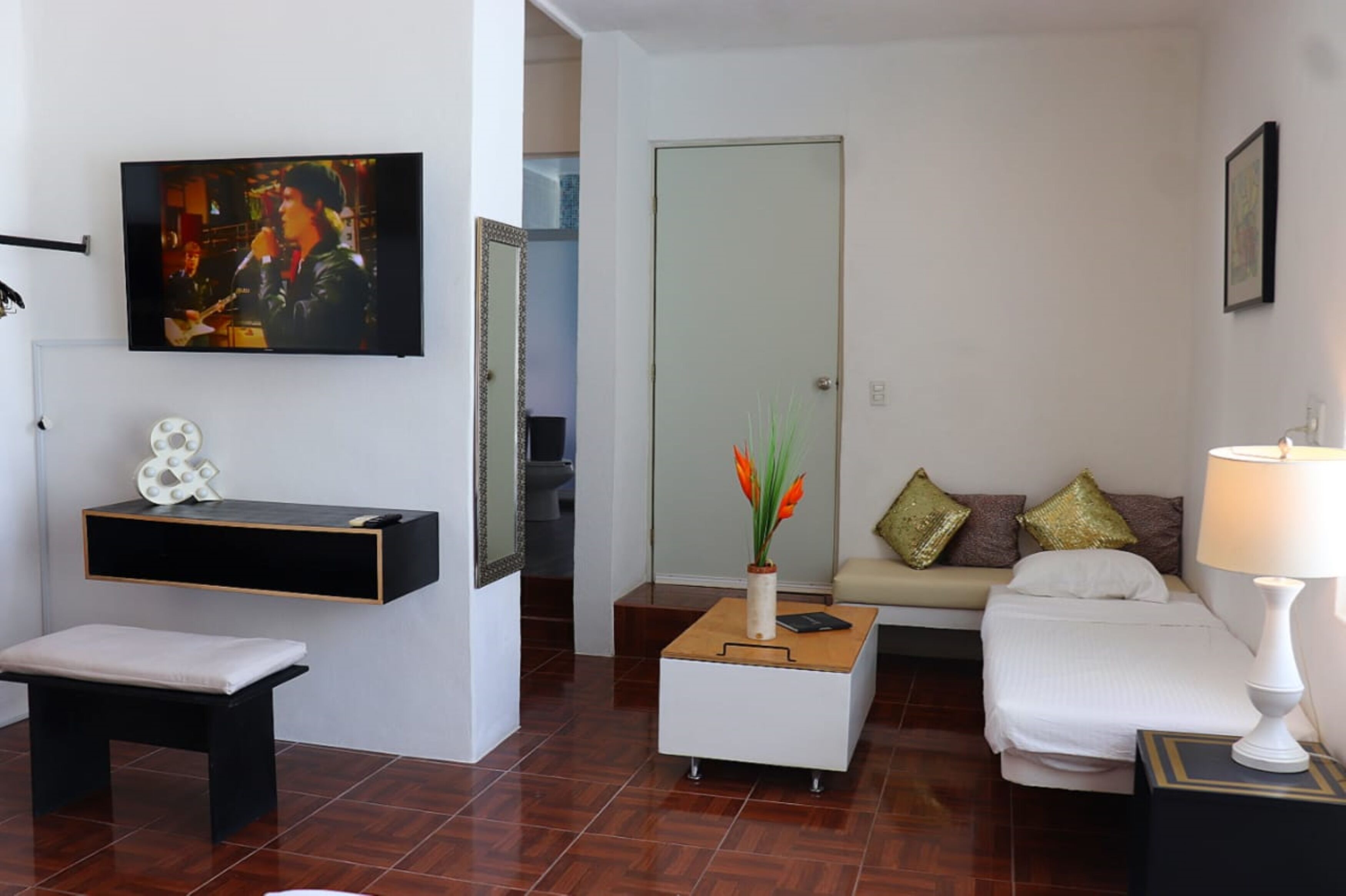 A modern living room with a flat-screen TV, a bench, a coffee table with a vase, a sofa with cushions, and a bedside table with a lamp.