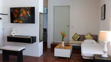 A modern living room with a flat-screen TV, a bench, a coffee table with a vase, a sofa with cushions, and a bedside table with a lamp.
