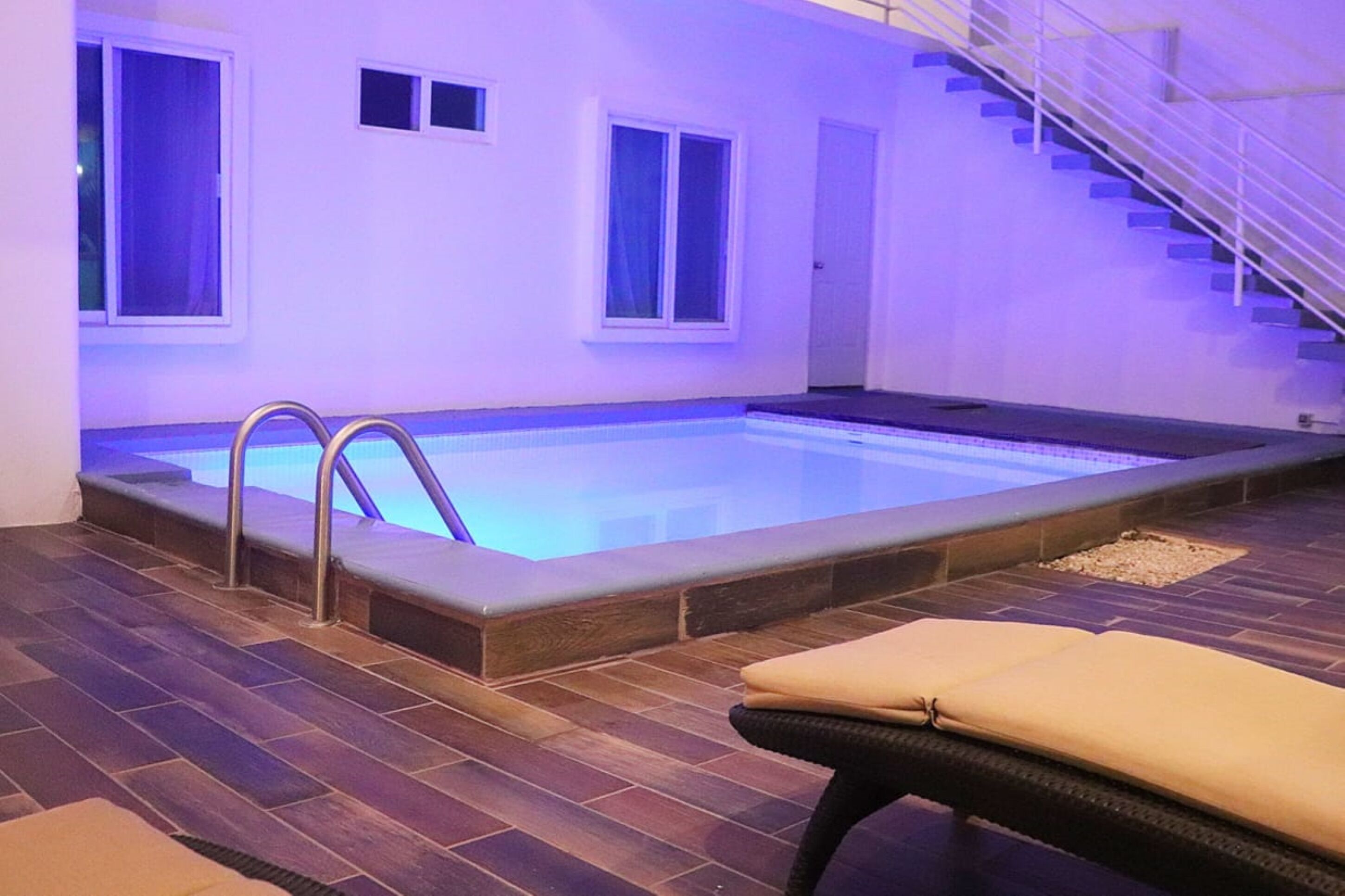 A swimming pool with a metal ladder, a cushioned lounge chair, and a staircase leading to an upper level.