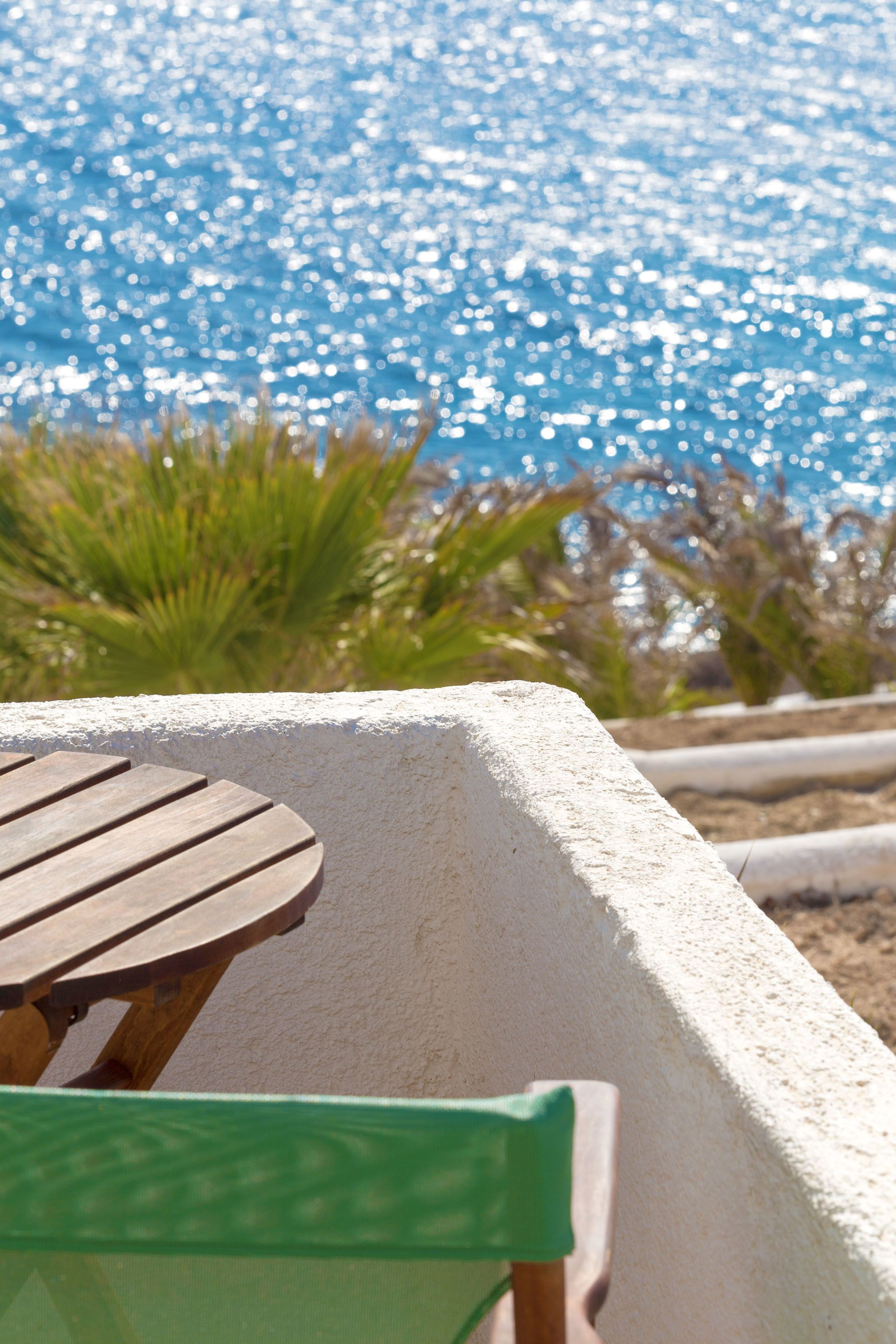 A wooden table and chair by a pool with a white wall and green plants in the background.