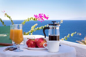 A table with two glasses of orange juice, a coffee pot, a plate with apples, and a white mug, set against a scenic view of the sea.