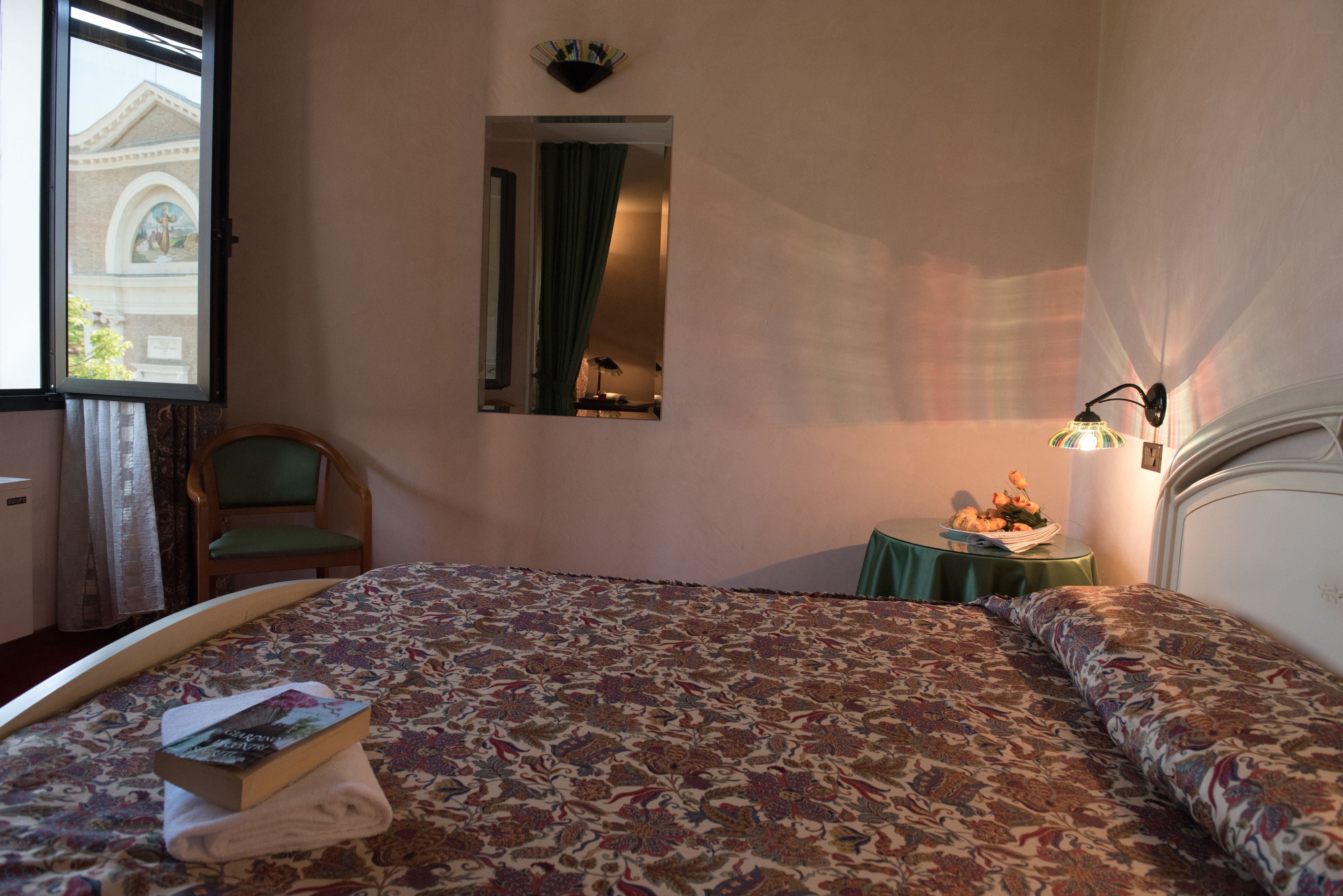 A hotel room with a large bed, a chair, a small table with flowers, and a window with a view of a building.