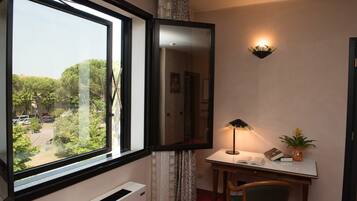 Ein Zimmer mit einem großen Fenster, einem Schreibtisch mit Lampe, einem Sessel und einer kleinen Pflanze.