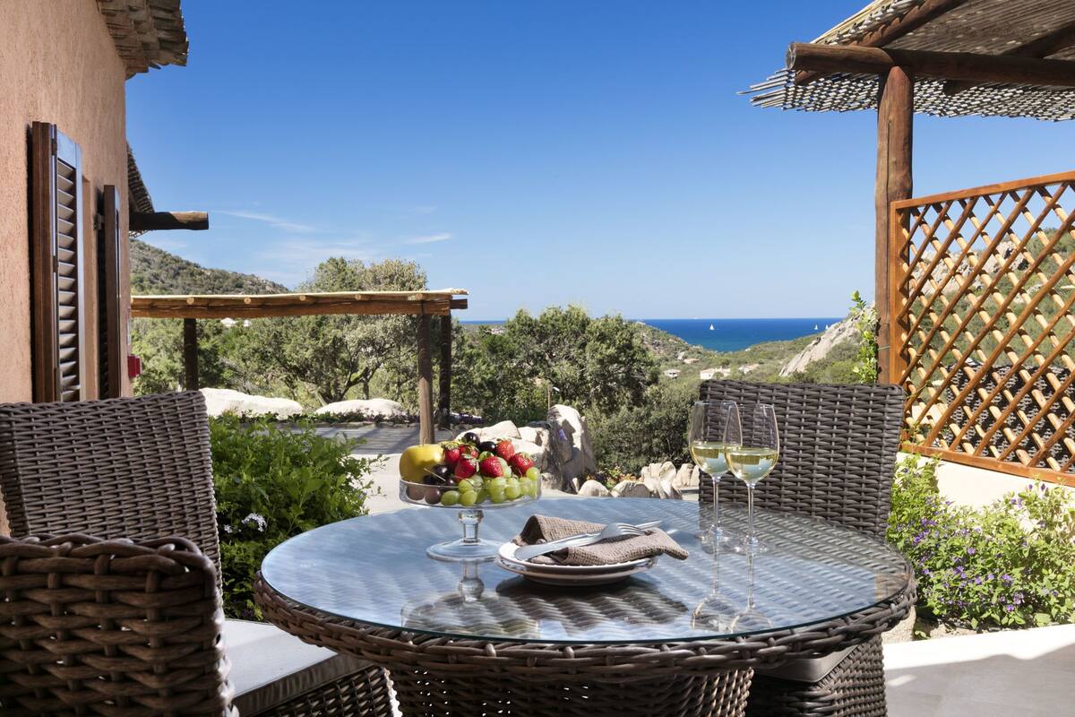 Een ronde glazen tafel met rieten stoelen, een fruitschaal en wijnglazen op een terras met uitzicht op een kustlandschap.