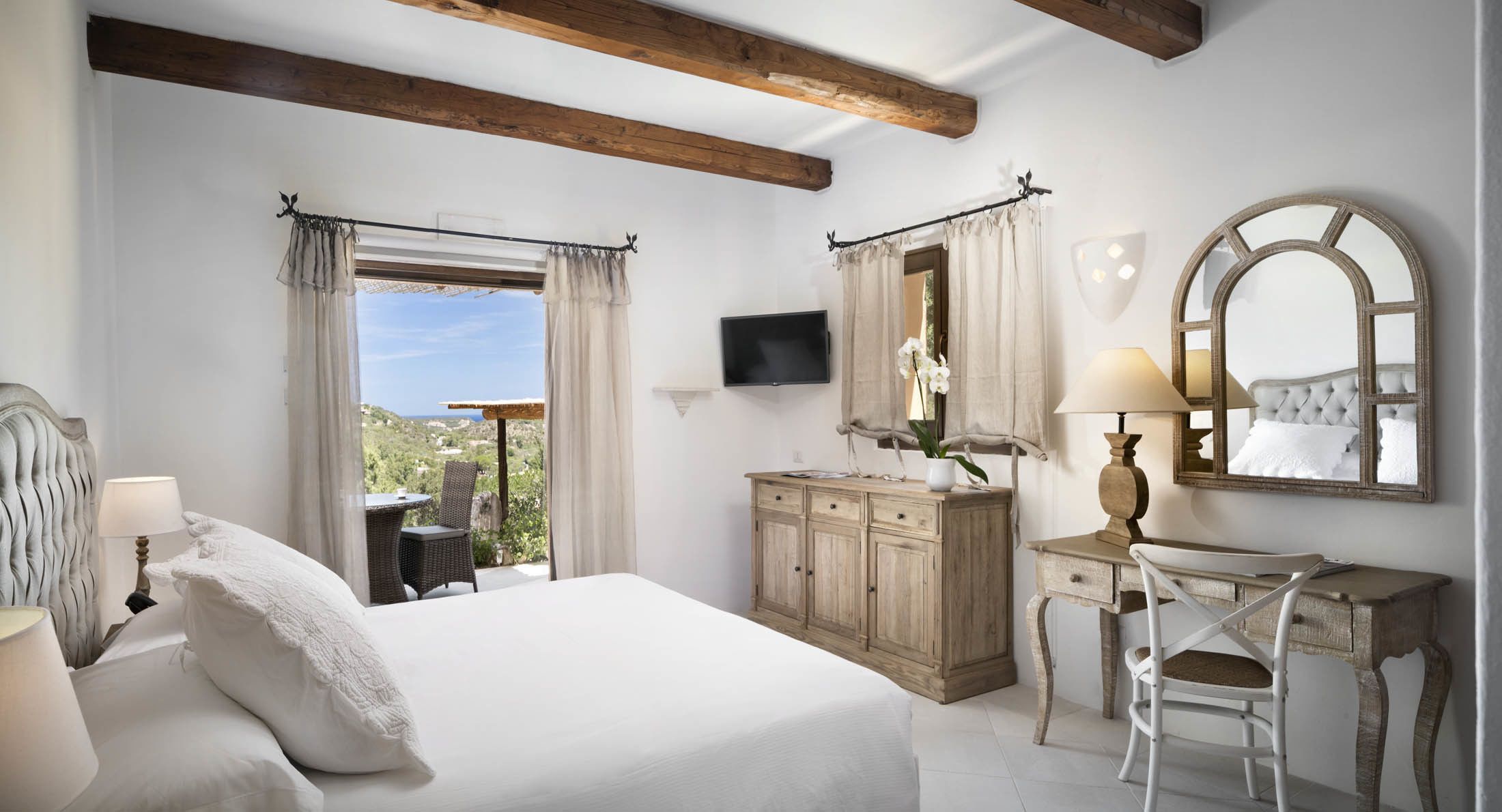 Quarto com cama, escrivaninha, cadeira, televisão e vista para o exterior.