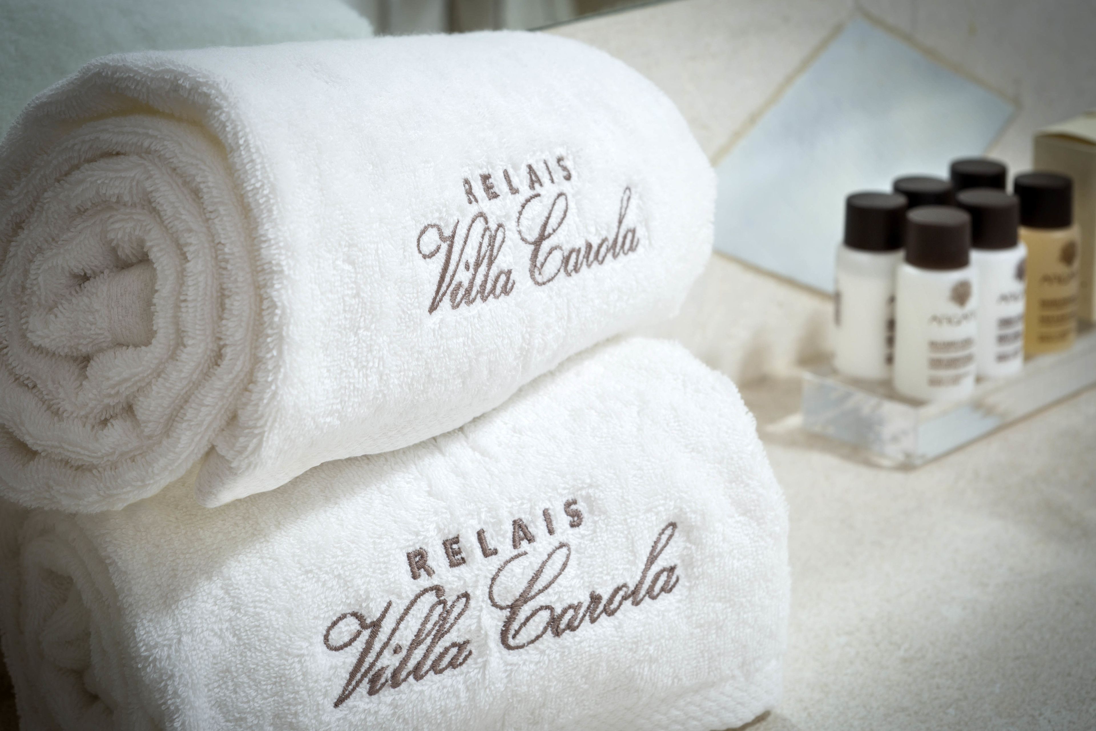 Dos toallas blancas enrolladas impecablemente con texto bordado, una bandeja con botellas y un fondo de azulejos.